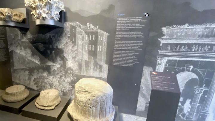Corridoio al piano terra di orientamento alla storia del castello: capitelli corinzi e rocchi di colonna esposti su basi, davanti a una grande immagine di sfondo in bianco e nero.