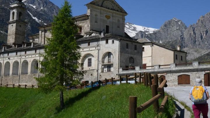 Esterno di santuario di San Magno a Castelmagno con loggiati, sfondo di montagne con neve, in visitatore a destra con zainetto in spalla