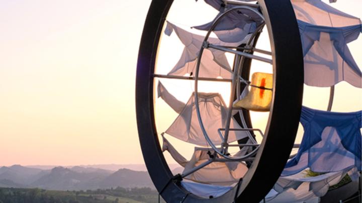 Particolare del banner della rassegna, con fotografia di opere d'arte contemporanea Foto di opera d'arta contemporanea composta da ruota in metallo con elementi di stoffa, sfondo di colline al tramonto