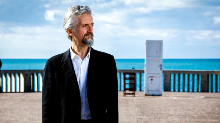 Il drammaturgo e attore Ascanio Celestini, dal sito https://sites.google.com/view/compagniadellozio Uomo con capelli e barba grigi, vestito di nero, sullo sfondo di passeggiata lungo il mare