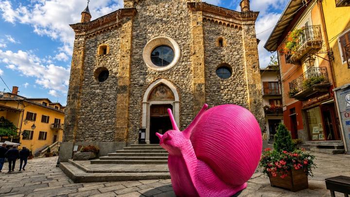 Fotografia da www.visitcuneese.it Facciata in pietra della chiesa parrocchiale di Limone Piemonte e davanti una scultura in plastica rosa rappresentante di Cracking Art una chiocciola gigante