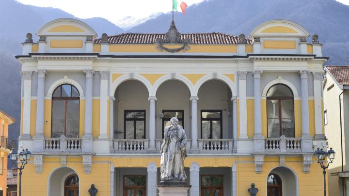 Facciate del teatro Civico di Varallo, da Wikipedia, fotografia di CIvvi Piazza con facciata di teatro ottocentesco in stile eclettico, in primo piano statua, sullo sfondo le Alpi