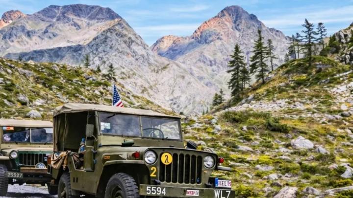 Particolare della locandina dell'evento, dal sito www.visitcuneese.it Fotografia: autoveicoli militari con bandiera statunitense percorrono strada di montagna, cime e conifere sullo sfondoa