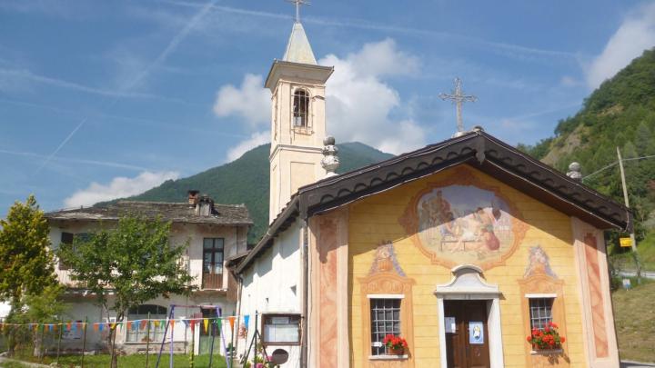 Cappella con facciata affrescata e campaniletto, bandierine decorative appese, sfondo di montagne