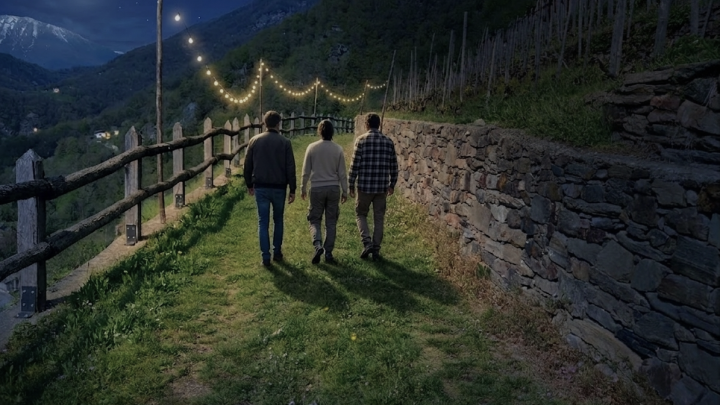 Tre persone camminano tra i vigneti di notte, con le montagne all'orizzonte.