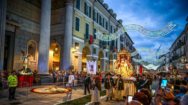 Fotografia da www.comune.cuneo.it Fotografia della processione serale della Madonna del Carmine in Via Roma a Cuneo: confratelli portano una statua dorata della Madonna fra due ali di folla su tappeti di petali di fiori con luminarie