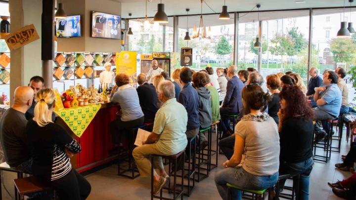 Immagine dal sito del Comune di Cuneo Show cooking all'Open Baladin di Cuneo: prodotti agricoli e gastronomici su un bancone in un ambiente con vetrate, con numeroso pubblico seduto su sgabelli